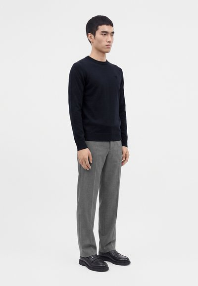 Jeune homme debout en pantalon gris, pull noir et chaussures noires sur un fond clair uni.