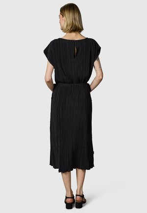 Kvinde med kort blondt hår iført en sort plisseret midi-kjole og sorte hælede sandaler, står med hænderne i lommerne og vender væk.