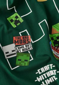 Grøn T-shirt med pixeliserede Minecraft karakterdesigns og tekst: "DE DÅRLIGE MOBBER," "KOMMER UD OM NATTEN," og "SKAB UDEN GRÆNSER."