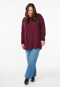 Burgunderfarbene langärmelige Bluse mit lässigem Schnitt, Rundhalsausschnitt und Seitenschlitzen, kombiniert mit hellblauen weiter geschnittenen Jeans und schwarzen Stiefeletten.