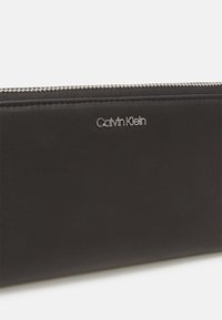 Calvin Klein black leather wallet featuring a sleek design, silver zipper, and embossed logo on the front. Smooth texture with minimal detailing.