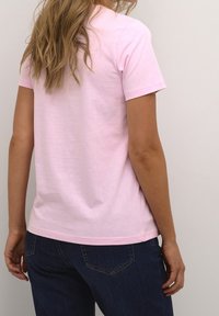 T-shirt en coton rose clair avec des manches courtes, col rond et coupe décontractée. La vue de dos met en avant la texture lisse et le design simple.