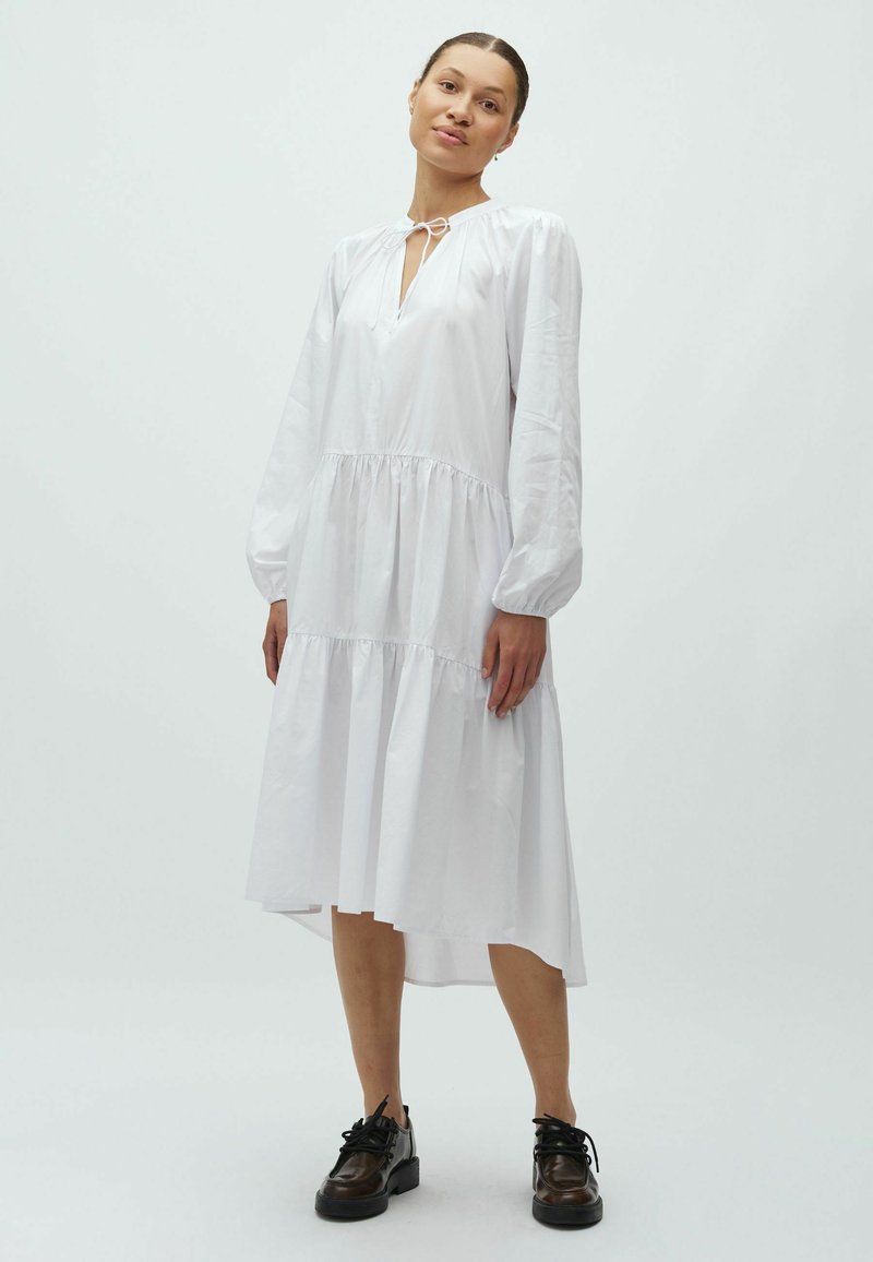 Robe blanche à manches longues avec une taille basse, une jupe à volants et un col à nouer. Portée avec des chaussures noires à lacets. Texture de tissu lisse.