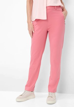 Kvinde iført lyserøde bukser med lige ben og lyse beige sneakers, stående mod en hvid baggrund med den ene hånd i lommen.