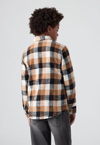 Camicia di flanella a quadri in arancione, nero e bianco. Presenta maniche lunghe, chiusura con bottoni e una vestibilità rilassata. Indossata con jeans grigi.