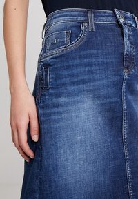 Denimhame keskisinisena, millel on sobiv vöökoht, kerge A-line kuju ja nähtavad õmblused. Eesmine külg on varustatud tasku detailidega.