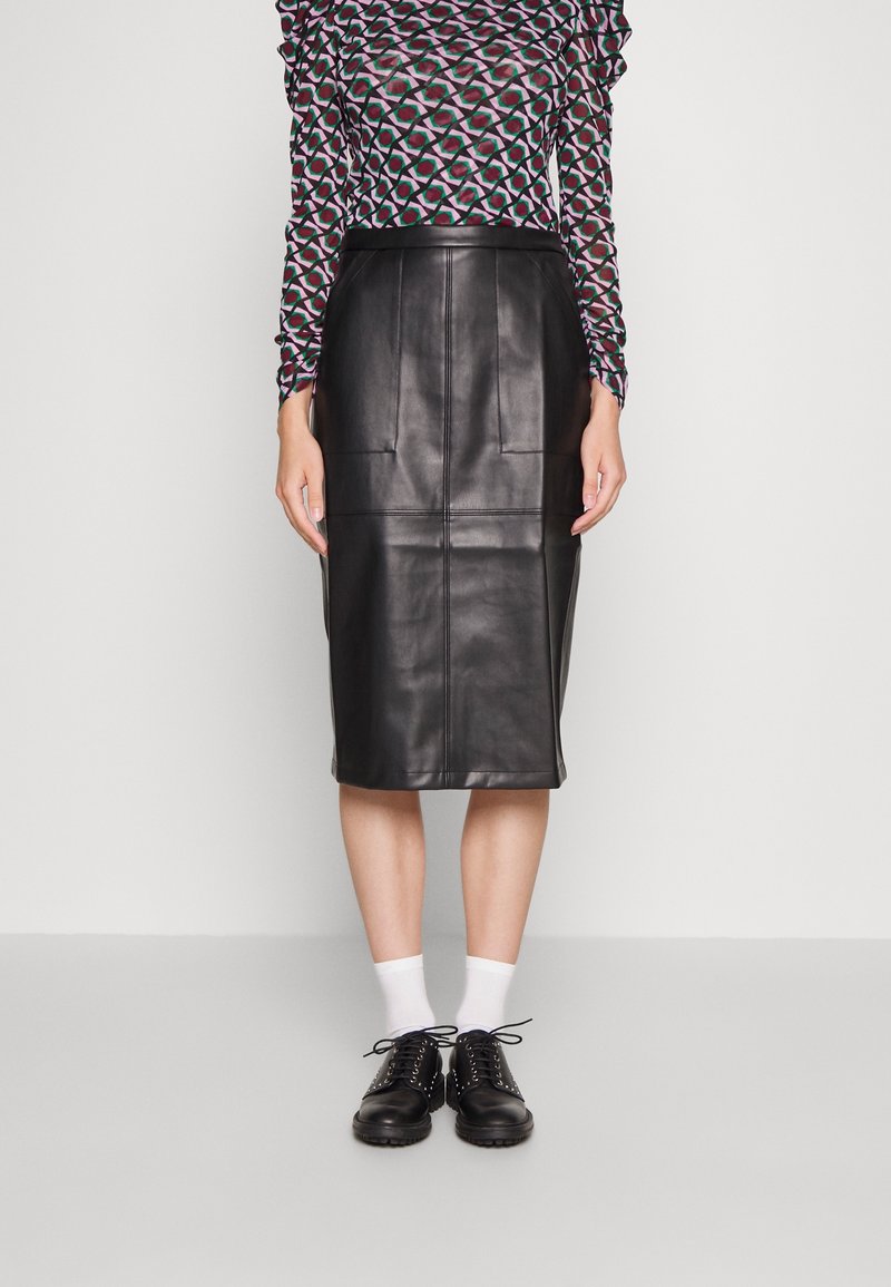 Femme portant une jupe en cuir noire longueur genou, un haut à manches longues à motifs, des chaussettes blanches et des chaussures noires à lacets, debout sur un sol gris.