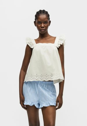 Žena nosi biele tričko bez rukávov s volánikovou ažúrovou lemovkou a svetlomodré pruhované šortky s volánikmi na lemoch.