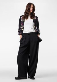 Woman wearing a black floral blazer over a white shirt, black wide-leg trousers, and black loafers, standing with hands in pockets.