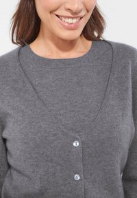 Frau lächelt, trägt eine graue Strickjacke mit weißen Knöpfen über einem passenden grauen Rundhalsoberteil.