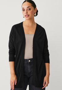 Zwarte cardigan met lange mouwen en een open voorkant, gemaakt van een zachte gebreide stof. Voorzien van geribde manchetten en een zoom, gelaagd over een beige top.