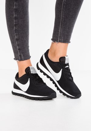 Černé tenisky s bílým logem Swoosh, mají strukturovaný svršek a polstrovanou podrážku, s viditelnou nášivkou loga na jazyku.