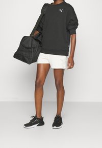 Personne portant un sweat-shirt Puma noir, un short Puma blanc, des baskets noires, et portant un sac de sport noir sur l'épaule.