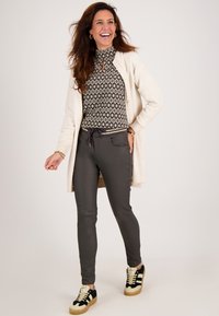 Lichtbeige cardigan over een zwart-witte gepatterneerde top met hoge hals. Grijze slim-fit broek en zwarte sneakers met crèmekleurige accenten. Glimlachende pose.