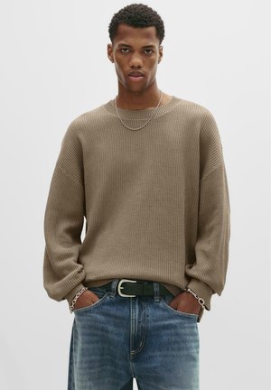 Jonge man met kort, krullend haar, gekleed in een beige wafelgebreid vest, zilveren ketting en armband, en losse blauwe jeans met een zwarte riem.