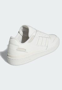 Sapatilha branca Adidas com design de cano baixo, três riscas laterais e sola texturizada, mostrada a partir de um ângulo traseiro sobre um fundo claro.