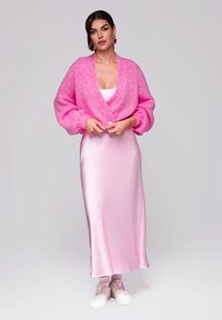 Růžová pletená kardigán s objemovými rukávy přes saténové růžové midi šaty, doplněné bílými teniskami.