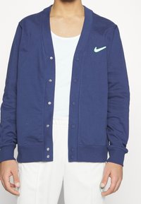 Man wearing navy blue button-up cardigan with a green Nike logo, white tank top, and white pants with an elastic waistband.