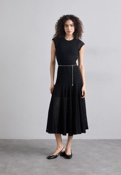 Schwarzes Kleid mit kurzen Ärmeln, strukturiertem Stoff, A-Linie Silhouette, detailliertem Saum und einer Kettengürtel. Kombiniert mit schwarzen Slingback-Schuhen.