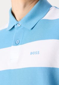 Polo rayé bleu clair et blanc avec col et deux boutons, avec le logo "BOSS" sur la poitrine.
