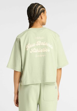 Femme avec des cheveux tressés portant un t-shirt cropped oversized vert clair de New Balance Athletics et un pantalon assorti avec poches arrière.