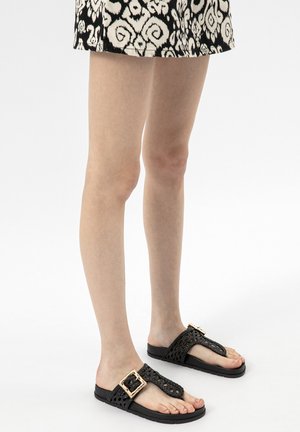 Jambes portant des sandales tissées noires avec de grandes boucles dorées et une jupe à motifs noir et blanc sur un fond uni.