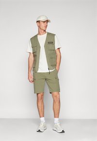 Gilet verde oliva con molte tasche sopra una maglietta bianca, abbinato a pantaloni corti coordinati; indossa un cappellino beige e sneakers bianche.