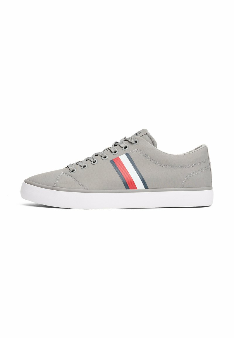 Sneaker in tela grigia con design a basse, caratterizzata da una suola in gomma bianca e accenti a strisce rossa, bianca e blu navy sui lati.