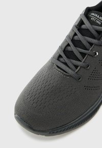 Black athletic shoe featuring a textured mesh upper, round laces, and rubber sole. Side has small ventilation holes and logo detail.