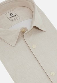 Camisa de botones color beige hecha de tela texturizada. Presenta un clásico cuello amplio y un patrón sutil. Dos botones cerca del cuello. Etiqueta de la marca visible.