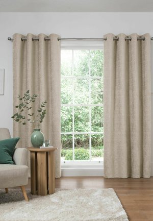 Beigefarbene Vorhänge rahmen ein großes Fenster mit sichtbarem grünem Laub draußen, neben einem beigen Sessel und einem hölzernen Beistelltisch mit grüner Vase.