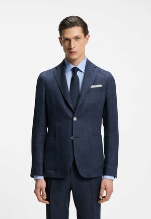 Uomo che indossa un abito blu scuro strutturato con camicia azzurra, cravatta blu navy a pois e fazzoletto bianco nel taschino, in piedi davanti a uno sfondo bianco uniforme.