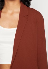 Blazer v rezavé barvě vyrobený z hladkého materiálu, s výstřihem do V a otevřeným předním dílem. Nositý přes bílý žebrovaný top.