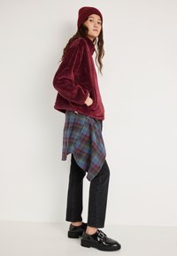 Bluza polarowa w bordowym kolorze, z teksturowanej tkaniny i wysokim kołnierzem. Połączona z niebiesko-zieloną koszulą w kratę oraz czarnymi spodniami o prostych nogawkach.