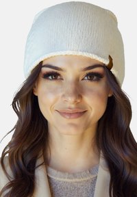 Młoda kobieta o długich brązowych włosach, ubrana w biały dziergany beret i sweter w neutralnych tonach, delikatnie uśmiechająca się na tle jednolitego tła.