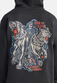 Twee felle honden aan een ketting, een skeletten conducteur, en een rennende cartoon hond op treinsporen met de tekst "Ride the Rails" en "Element & Timber" op een zwarte hoodie.
