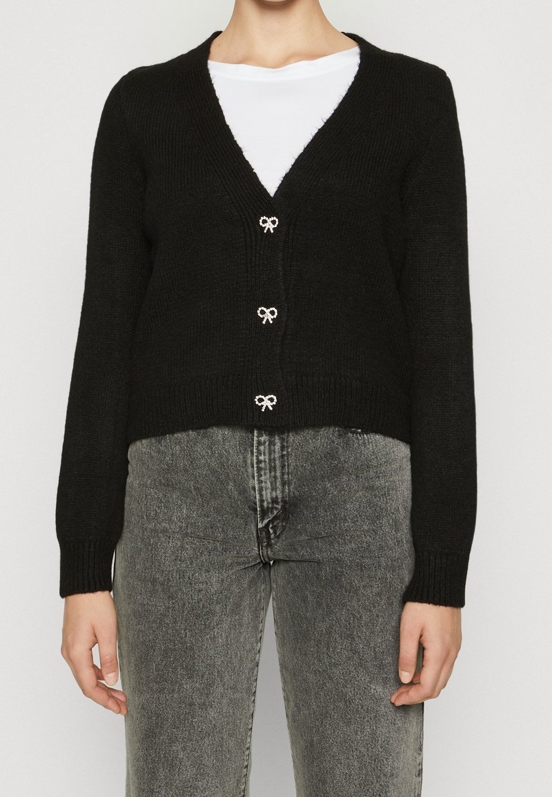 Cardigan noir court en maille texturée, avec un col en V, trois boutons décoratifs en forme de nœud et des poignets côtelés, associé à un jean gris.