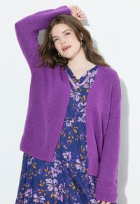 Frau posiert mit der Hand über dem Kopf, trägt eine lila Strickjacke über einem blauen Blumenkleid mit orangefarbenen und lavendelfarbenen Akzenten.