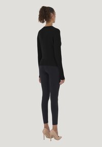 Sort strikket sweater med lange ærmer og en tætsiddende kant, kombineret med sorte leggings og nøgenfarvede højhælede sko, set bagfra.