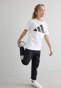 Balta T-kreklu ar lielu melnu ģeometrisku logo, melnas sporta bikses ar baltām strīpām un baltām sporta apaviem.