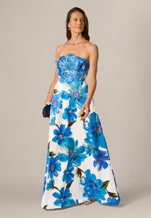 Vrouw in strapless witte jurk met grote blauwe bloemenprint en versierde blauwe bloemdetail op het lijfje, houdend een kleine donkere clutch.