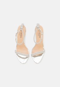 Hopeasäihkeät sandaalit, joissa on kiiltävä metallipinta ja rhinestone-koriste nauhassa. Pehmustettu vaaleanbeige sisäpohja, avoin kärkilinja.