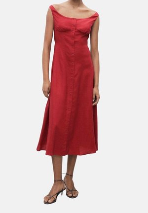 Femme portant une robe midi rouge sans manches, épaules dénudées, boutonnée sur le devant, avec des sandales noires à talons hauts et fines bretelles sur un fond blanc.