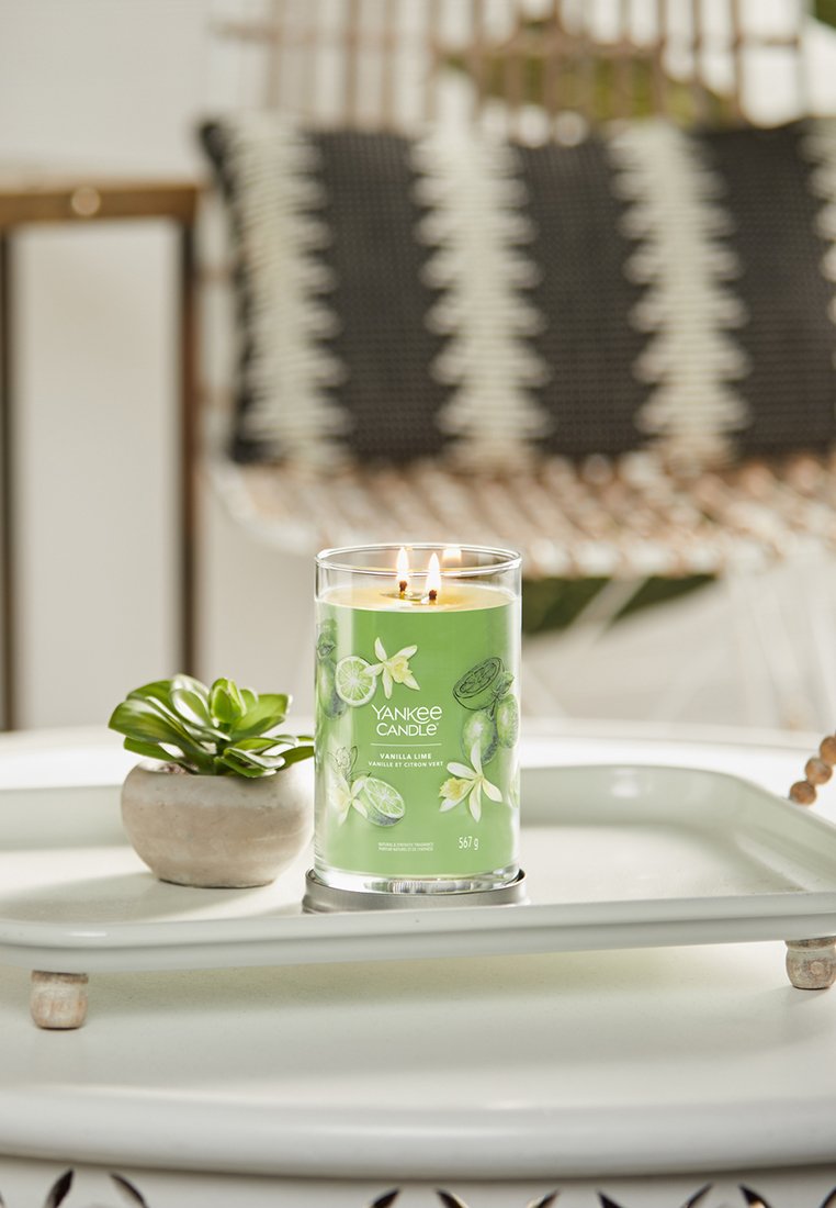 Yankee Candle in einem durchsichtigen Glasbehälter mit grünem Etikett, das Illustrationen von Vanille und Limette zeigt, mit zwei Dochten, auf einem weißen Tablett liegend.