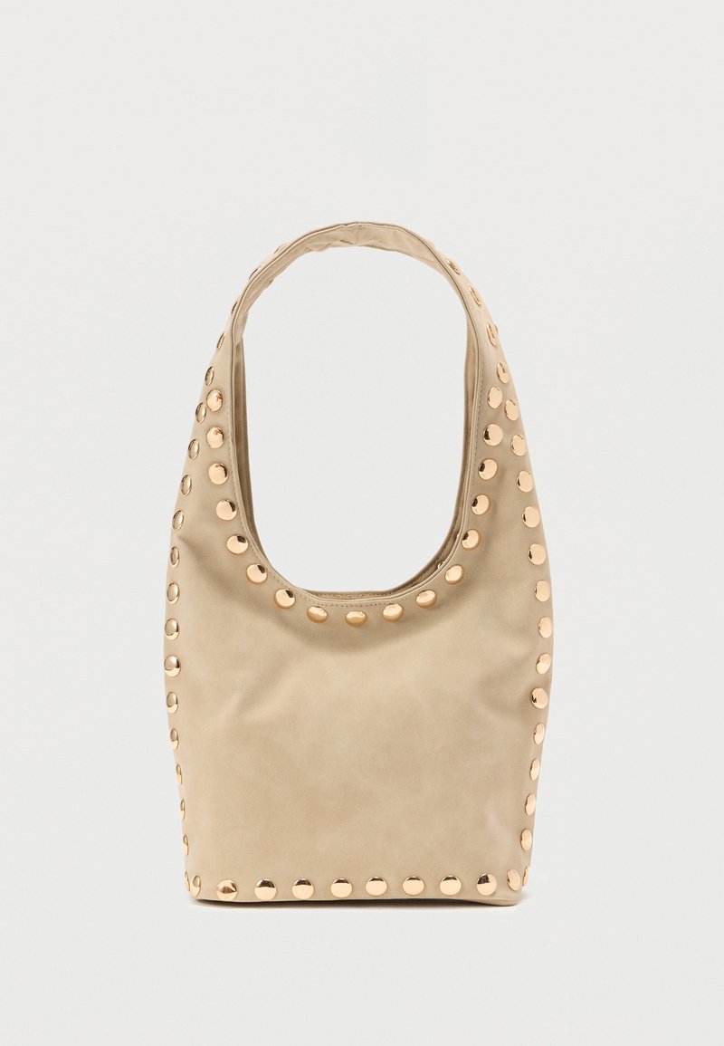 Sac à main beige de forme arrondie, texture lisse et clous circulaires dorés autour des bords. Anse unique et ouverture spacieuse.