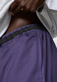 Violeta sporta bikses ar melnu elastīgu jostu, ar teksturētu, perforētu audumu un sānu kabatām.