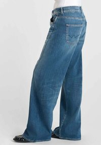 Jean en denim à coupe large avec un lavage bleu clair, présentant une taille haute, un design classique à cinq poches et une couture de marque sur la poche arrière.