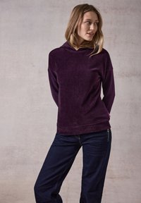Top cappuccio rigato viola abbinato a pantaloni in denim scuro. Il tessuto ha un aspetto morbido e testurizzato. Dettagli notevoli includono una cerniera laterale.
