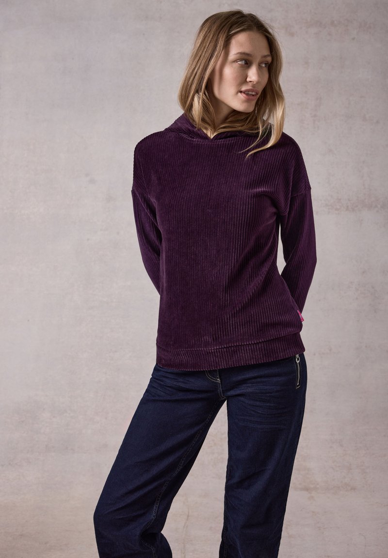 Top cappuccio rigato viola abbinato a pantaloni in denim scuro. Il tessuto ha un aspetto morbido e testurizzato. Dettagli notevoli includono una cerniera laterale.