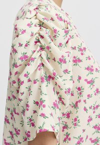Květinová blůza v krémové barvě s nabíranými rukávy. Má růžové květiny a zelené listy. Vyrobena z lehkého materiálu s jemnou texturou.